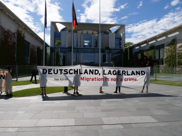 Eine Gruppe von Menschen hält ein Transparent mit der Aufschrift 'Deutschland, Lagerland Migration ist kein Verbrechen' vor einem Gebäude mit Fenstern, umgeben von Gras, einem Metallzaun, Pflanzen, Bäumen und Fahnenstangen.
