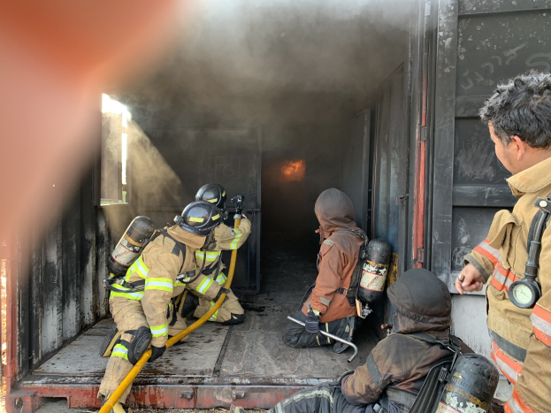 Feuerwehrleute in Helmen und Sauerstoffflaschen arbeiten von der Rückseite eines raucherfüllten Trucks an einem Brandort, wobei einer einen Schlauch hält.