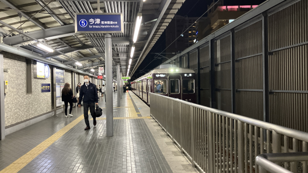 Ein Zug fährt auf ein Bahnsteiggleis zu, mit Menschen in der Nähe, einem Geländer auf der rechten Seite, Gebäuden im Hintergrund, Bahnsteigbeleuchtung und einer Anzeigetafel mit Text.