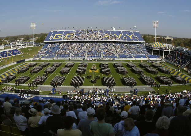 Eine große Menge schaut einem Fussballspiel in einem grünen Stadion unter einem klaren blauen Himmel zu, mit Laternen, Bäumen und Gebäuden im Hintergrund.