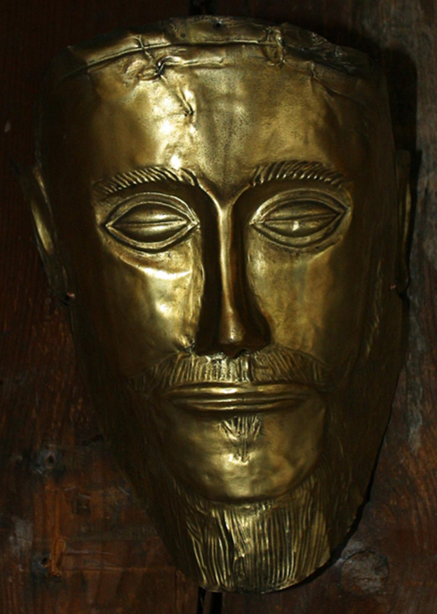 Eine goldene Maske von Jesus Christus mit detaillierten Verzierungen, Bart, Heiligenschein, geschlossenen Augen, leicht geöffnetem Mund, weißem Gewand und Dornenkrone, die an einer hölzernen Wand hängt.