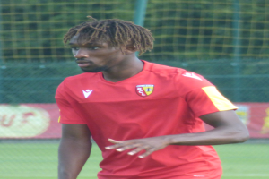 Ein Mann mit Dreadlocks steht auf einem Fußballfeld und trägt ein rotes T-Shirt und Shorts, im Hintergrund gibt es ein Netz und eine Tafel mit der Aufschrift "RCCL - Ghanaian Premier League"