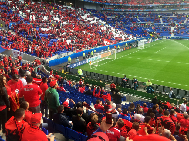 Ein vollbesetztes Stadion mit sitzenden und stehenden Zuschauern, Fahnen, einem Torpfosten und Deckenleuchten, während die Menge gespannt auf den Beginn des Spiels wartet.
