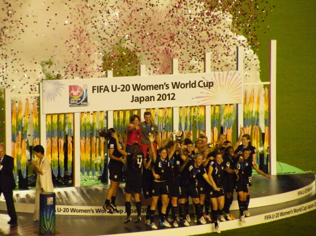 Schwarztrikotierte Frauenfußballspielerinnen auf einer Bühne, die einen Sieg feiern, umgeben von einer Menge mit Kameras, mit einem Hintergrundboard und einem grasbedeckten Boden.