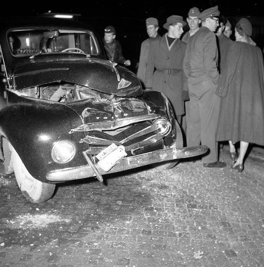 Schwarzes und weißes Bild eines schwer beschädigten Autos am Straßenrand mit eingedellter Motorhaube, zersplitterten Fenstern und einer Gruppe von Menschen, die es besorgt untersuchen.