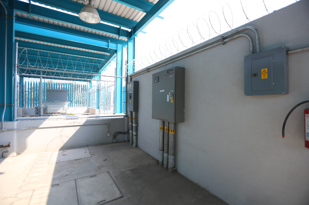 Ein Raum mit einem Feuerlöscher auf der rechten Seite, elektrischen Schächten und Rohren an der Wand, einer Deckenleuchte und einem Zaun im Hintergrund, wahrscheinlich eine elektrische Umspannstation.