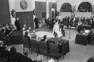 Schwarz-weißes Foto einer Hochzeitszeremonie in einem großen Raum mit sitzenden und stehenden Gästen, Tischen mit Papieren und Mikrofonen, einer Rückwand mit Rahmen, Blumenbouquets, einem Vorhang und einem Podium mit Mikrofonständer.