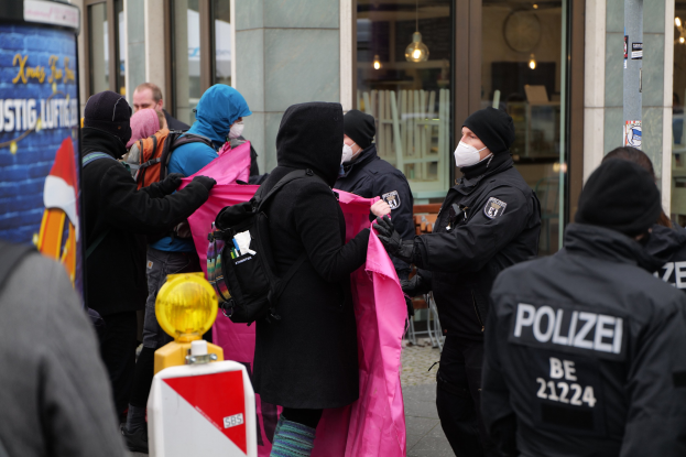 Gruppe von Menschen vor einem gläsernen Gebäude mit Deckenbeleuchtung, einige halten Kleidung oder tragen Mützen und Taschen, ein Polizist verhaftet eine Frau, mit einem Pfahlschild und einem Wasserzeichen.