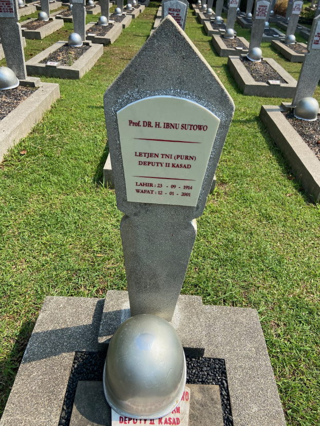 Ein Friedhof mit zahlreichen Grabsteinen, die mit Metallhelmen gekrönt sind, Rasenboden und Bäumen im Hintergrund.