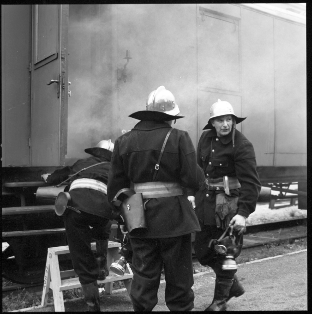 Feuerwehrmänner mit Helmen auf einem Bahngleis, einer sitzt auf einem Hocker, mit einem rauchenden Zug im Hintergrund.