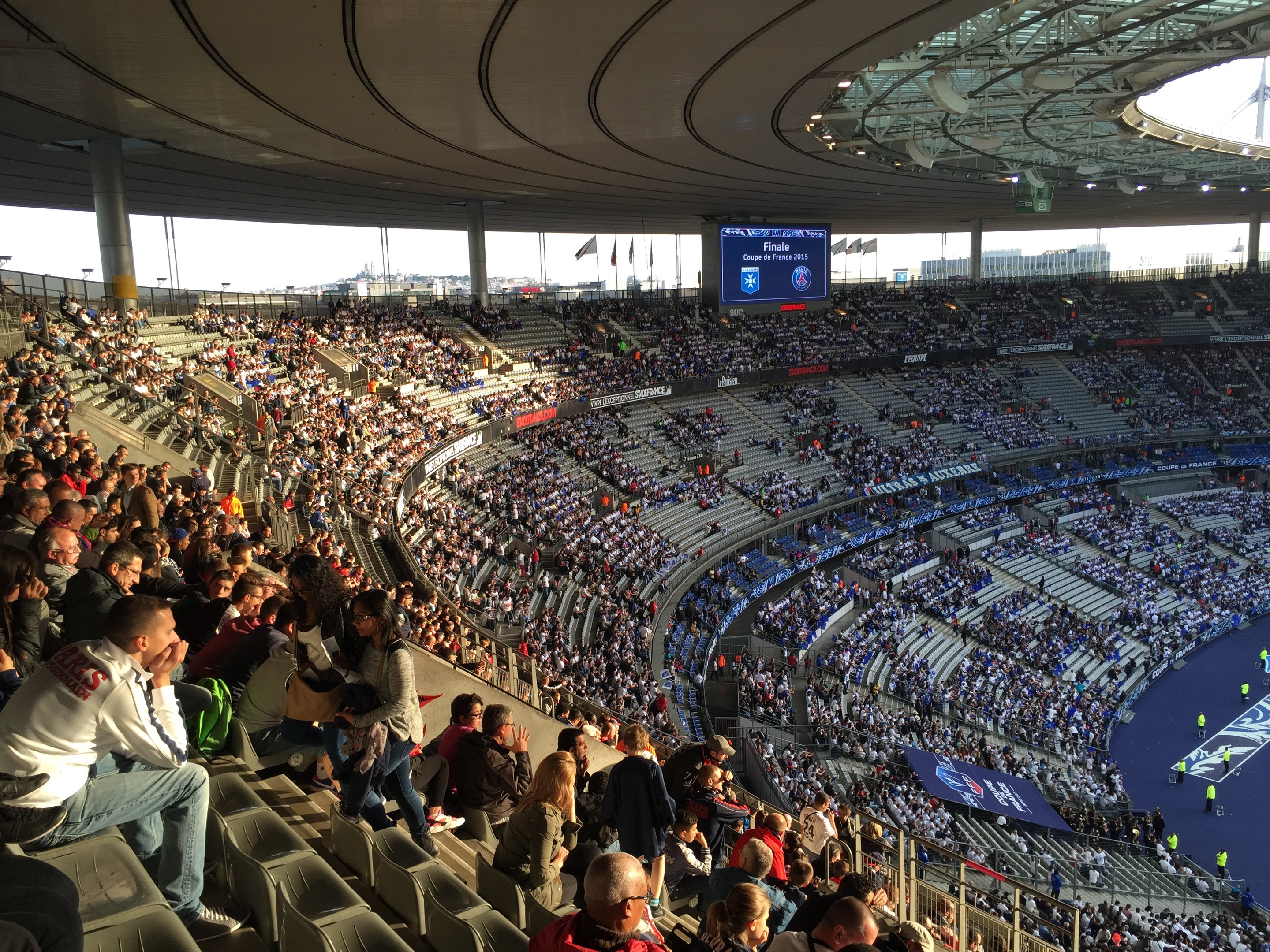 Große Zuschauermenge in einem Stadion bei einem Fußballspiel mit einer Bühne, Fahnen und einem Bildschirm im Hintergrund, identifiziert als Allianz Arena in München, Deutschland.