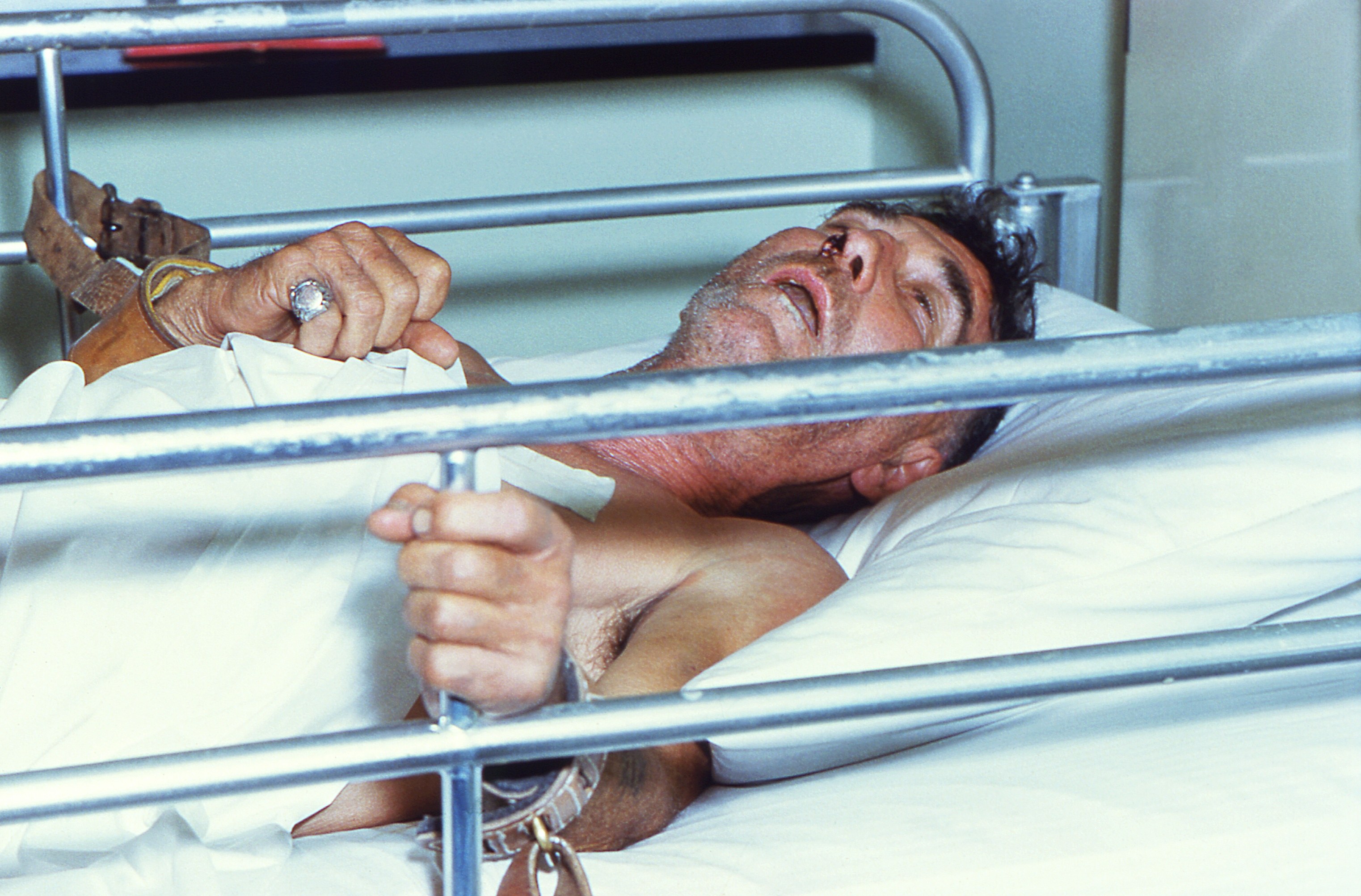 Ein Mann liegt in einem Krankenhausbett, die Hände auf dem Kopf, mit Metallstangen in der Nähe und einer Wand im Hintergrund, der besorgt mit geschlossenen Augen und zusammengesunkener Haltung appears.