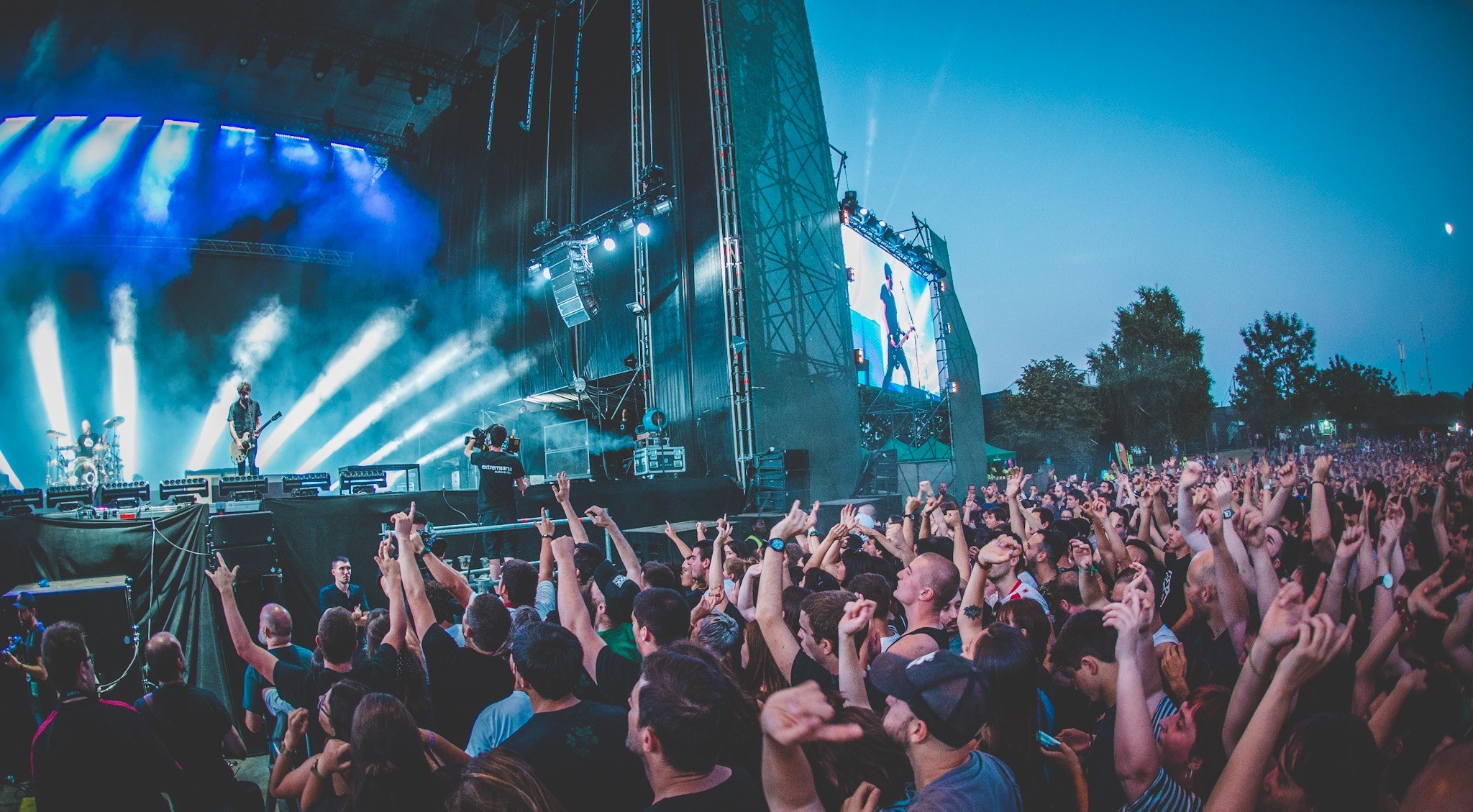 Eine Menschenmenge auf einem Musikfestival mit erhobenen Händen vor einer Bühne mit Instrumenten, Lichtern und einem Bildschirm, vor einem Wald und einem klaren blauen Himmel.