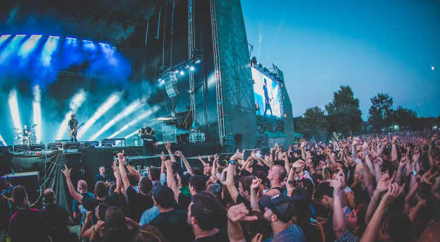 Eine Menschenmenge auf einem Musikfestival mit erhobenen Händen vor einer Bühne mit Instrumenten, Lichtern und einem Bildschirm, vor einem Wald und einem klaren blauen Himmel.