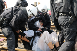 Eine Gruppe von Polizisten in Einsatzausrüstung verhaftet einen Mann in einem weißen Kleid auf einem Bahnsteig, andere sind im Hintergrund sichtbar.