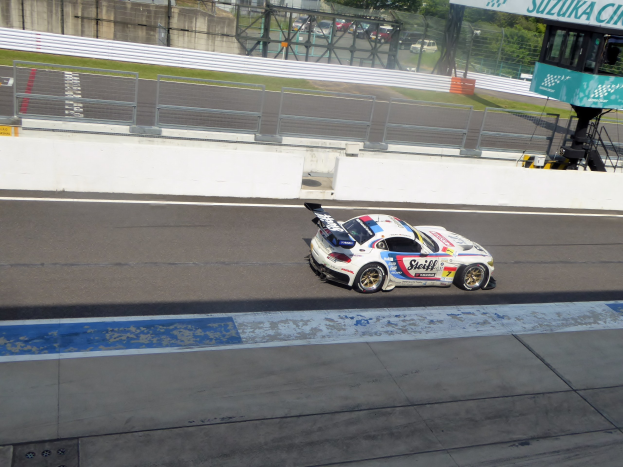 Ein Porsche 911 GT3 RSR auf einer Rennstrecke während der 24 Stunden von Le Mans, mit einem Zaun, Gras, Bäumen, Gebäuden und einem klaren blauen Himmel im Hintergrund und einer Tafel auf der rechten Seite.