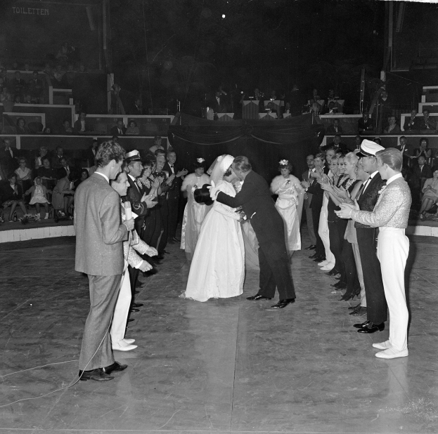 Ein Schwarz-Weiß-Foto eines Paares, das sich auf der Bühne umarmt und küsst, während es von klatschenden Menschen umgeben ist.