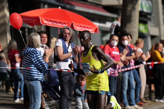 Ein Mann in einem gelben T-Shirt und Laufschuhen sprintet durch einen Marathon, während eine Menschenmenge hinter ihm jubelt, mit einem Schirm und einem Kinderwagen im Hintergrund.