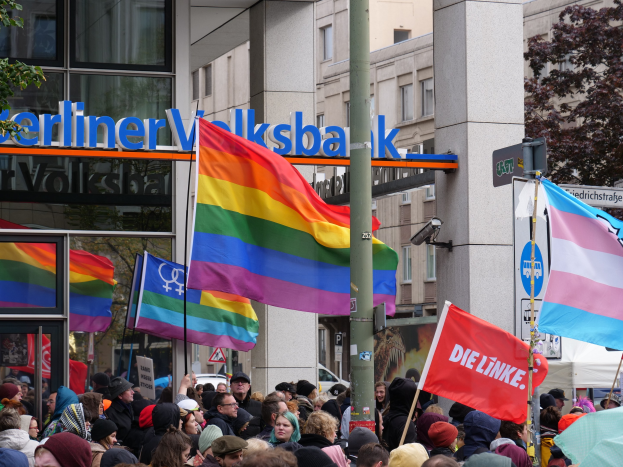 Große Gruppe von Menschen mit Fahnen und Transparenten vor einem Gebäude während einer Christopher Street Parade in Berlin, mit Bäumen und einem Mast im Vordergrund und Gebäuden im Hintergrund.