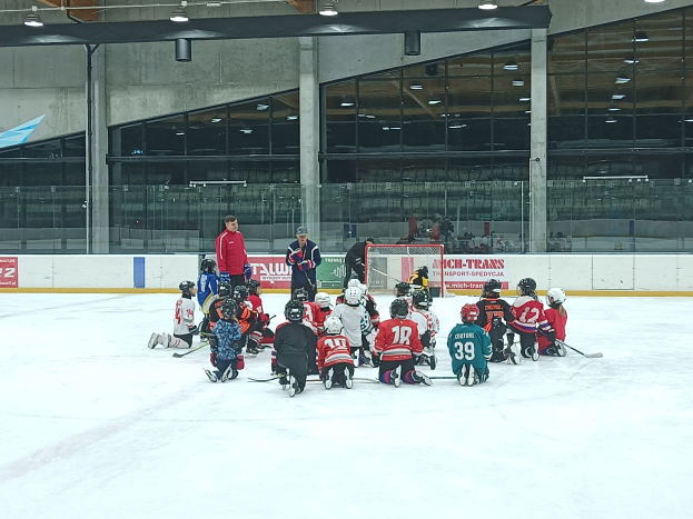 Eine Gruppe von Kindern in Helmen und mit Hockey-Schlägern sitzt auf einem Eisstadion, mit Glaswänden, Säulen, Deckenleuchten und Texttafeln im Hintergrund.