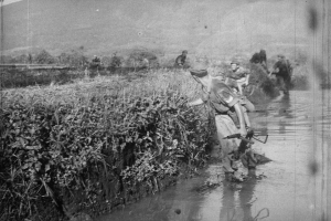 Schwarz-weißes Bild von Männern, die durch einen Fluss gehen, einer hält eine Waffe, mit Pflanzen auf der linken Seite und Hügeln im Hintergrund unter einem klaren Himmel.