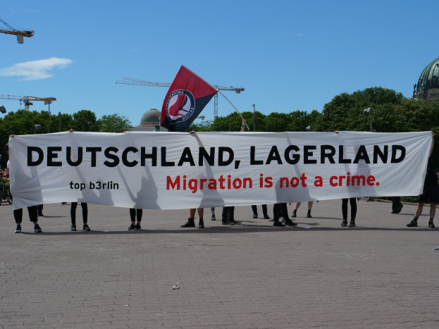 Eine Gruppe von Menschen, die ein Banner mit der Aufschrift "Deutschland, Lagerland Migration ist kein Verbrechen" und eine Flagge halten und auf dem Boden mit Straßenlaternen, Straßenlaternen, Bäumen, Kränen, einem Gebäude und einem bewölkten Himmel im Hintergrund stehen.