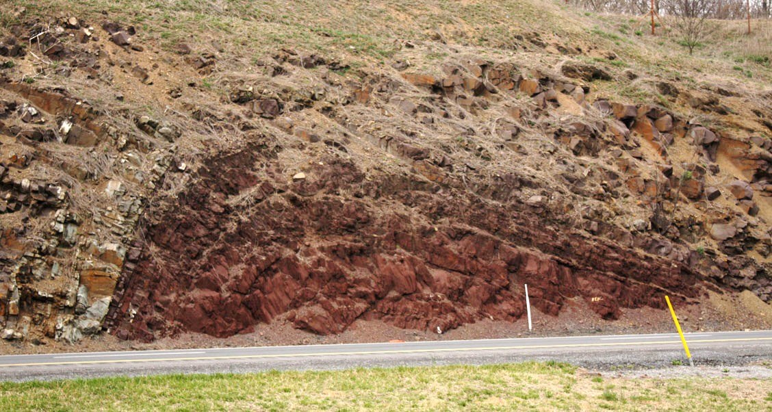 Großer Erdrutsch an der Seite einer Straße, mit Gras unten, einem Hügel in der Mitte, Bäumen und Himmel im Hintergrund.