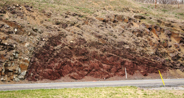 Großer Erdrutsch an der Seite einer Straße, mit Gras unten, einem Hügel in der Mitte, Bäumen und Himmel im Hintergrund.