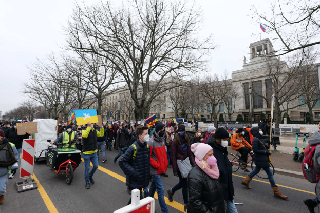 Eine große Protestdemonstration mit Menschen, die eine Straße in Washington, D.C. entlanggehen, einige halten Schilder und andere fahren Fahrräder, mit Bäumen und einem klaren blauen Himmel im Hintergrund.