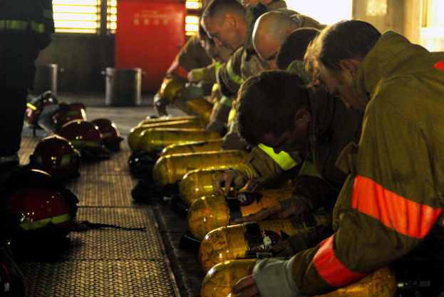Feuerwehrleute in Schutzausrüstung bei der Arbeit an einem Feuerwehrauto mit Ausrüstung und Helmen auf dem Boden, in einem Raum mit Fenstern, einer Tür und einem Müllcontainer.