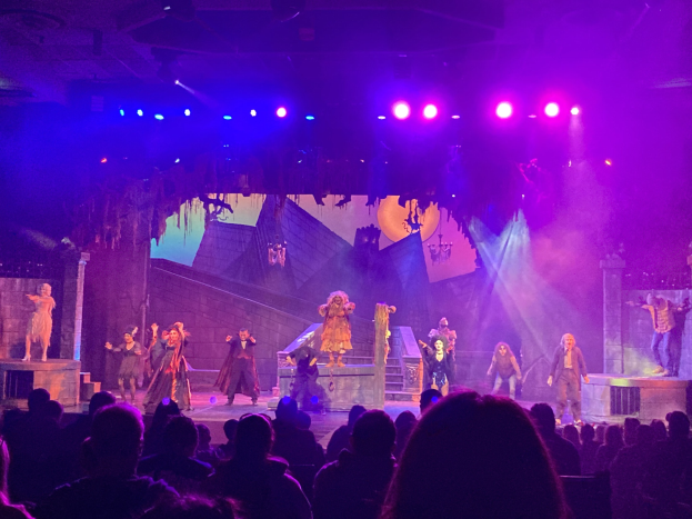 Eine Gruppe von Menschen in Kostümen steht auf einer hell erleuchteten Bühne, während das Publikum darunter auf Stühlen sitzt und eine Wand im Hintergrund zu sehen ist.