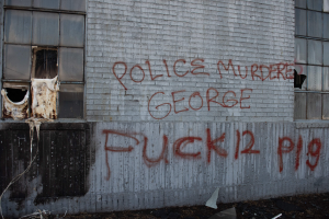 Graffiti an einer Gebäudewand mit der Aufschrift "Polizeimord George Fuck 12 P19" und Fenstern sowie verstreuten Gegenständen davor