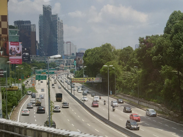 Eine belebte Autobahn mit hohen Gebäuden, Straßenlaternen, Schildern, Werbetafeln, Bäumen und einem bewölkten Himmel.