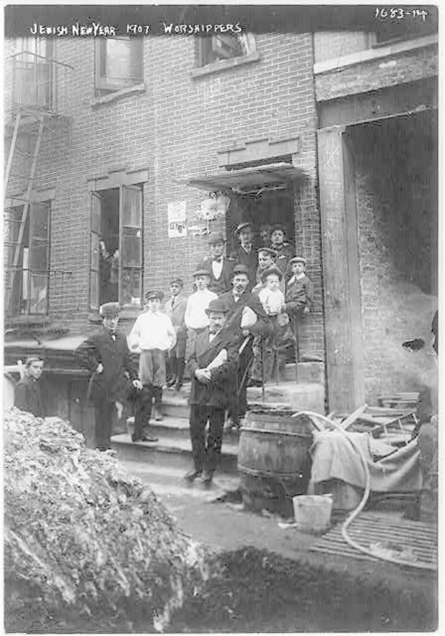 Schwarz-weiß-Fotografie von jüdischen Neujahrsfeiernden vor einem Gebäude mit Treppe in Brooklyn, New York, 1907, mit einem Haufen Erde links und hölzernen Gegenständen, einem Eimer und einem Rohr rechts.