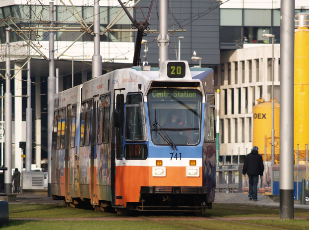 Blauer und oranger Tram auf einer Stadtstraße mit einem Fahrgast drinnen, eine Person zu Fuß auf dem Gehweg rechts und Gebäude, Masten und Drähte im Hintergrund.