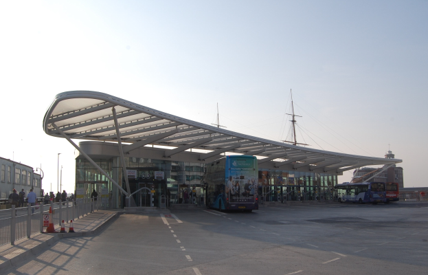 Ein Bushaltestelle mit parkenden Bussen, Menschen, die gehen, ein Geländer auf der linken Seite, Gebäude, Pfosten und Himmel im Hintergrund.