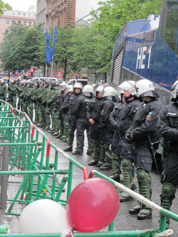 Polizeibeamte in Helmen und Uniformen stehen hinter einer Barriere, halten Waffen und haben Schienen, Luftballons, Bäume, Gebäude, Flaggen und einen klaren Himmel im Hintergrund.