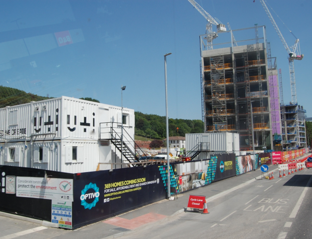 Eine Baustelle mit einem großen Gebäude im Bau, umgeben von Kränen, Straßenlaternen, Straßenschildern, Werbetafeln, Treppen, Geländern, Bäumen und einem klaren blauen Himmel.