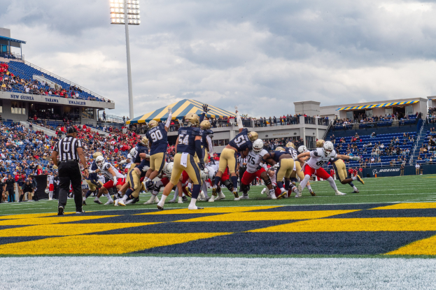 Fußballspieler in Helmen und Sportbekleidung konkurrieren in einem Stadion mit Zuschauern, Stühlen, Geländern, Beschriftungen, Laternenmasten und einem bewölkten Himmel im Hintergrund.