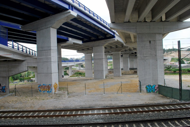 Eine Bahnschiene mit Graffiti an der Seite, die unter einer Brücke mit Pfeilern und Geländern hindurchführt, neben Pfählen mit Drähten, vor Bäumen, Gebäuden und einem klaren blauen Himmel.