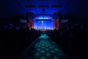 Ein großer Auditorium mit sitzenden Menschen, eine Bühne mit Musikern, die Instrumente spielen, und eine Decke mit Lichtern und Fernsehgeräten, was auf einen Konzertsaal für die Nationale Konferenz 2019 hindeutet.