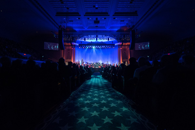 Ein großer Auditorium mit sitzenden Menschen, eine Bühne mit Musikern, die Instrumente spielen, und eine Decke mit Lichtern und Fernsehgeräten, was auf einen Konzertsaal für die Nationale Konferenz 2019 hindeutet.