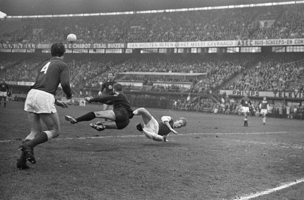 Schwarzes und weißes Foto von Männern, die Fußball in einem Stadion mit Zuschauern auf den Rängen und Transparenten im Hintergrund spielen.