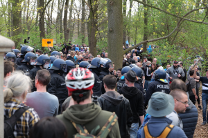 Große Menschenmenge vor einer Reihe von Helmpolizisten mit Ausrüstung, eine Säule links, Bäume im Hintergrund und einige Personen mit Kameras in einer Outdoor-Situation.