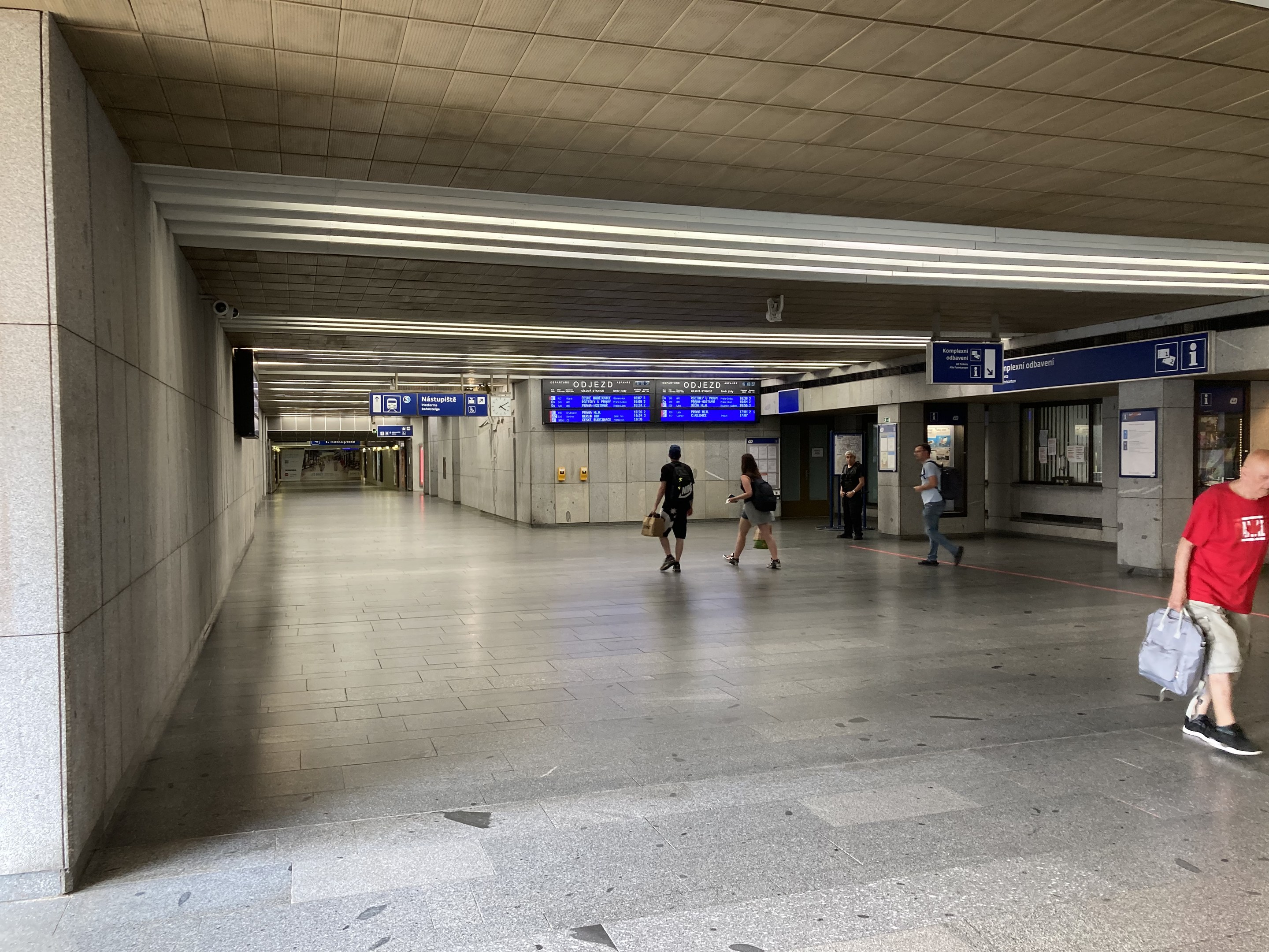 Menschen gehen durch eine U-Bahn-Station mit Texttafeln, Säulen und Deckenleuchten, einige tragen Taschen, mit einer Person in Sneakers in der Mitte.