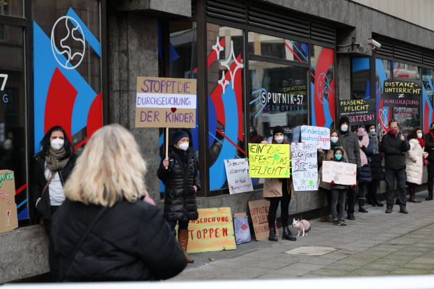 Menschen mit Masken, die Schilder und Plakate mit Text und Logos vor einem Gebäude mit Glaswänden und -türen halten, mit Kameras und einem Hund vor Ort, protestieren gegen eine deutsche Regierungspolitik.