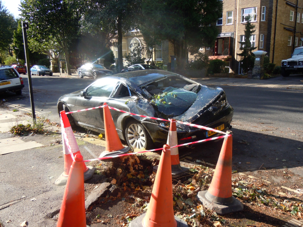 Ein besch├Ądigtwagen am Stra├čenrand mit orangen Verkehrskegeln, B├Ąumen, Geb├Ąuden und einem klaren blauen Himmel im Hintergrund.