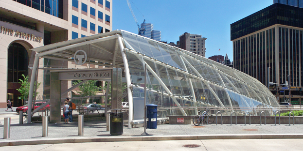 Eine belebte Stadtstraße mit hohen Wolkenkratzern, ein Bushaltestelle mit Glasdach, Menschen, Fahrzeuge, ein Metallzaun, ein Mülleimer, Pfosten, ein Schild, eine Ampel, Bäume und ein bewölkter Himmel.
