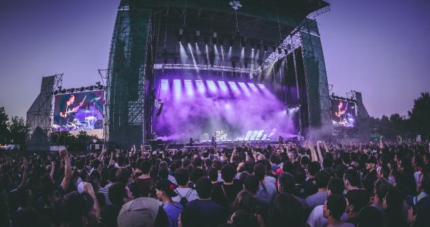 Eine große Menschenmenge auf einem Musikfestival schaut Künstlern auf einer hell erleuchteten Bühne mit Instrumenten und Bildschirmen zu, die vor einem Gebäude, Bäumen und einem klaren blauen Himmel aufgebaut ist.