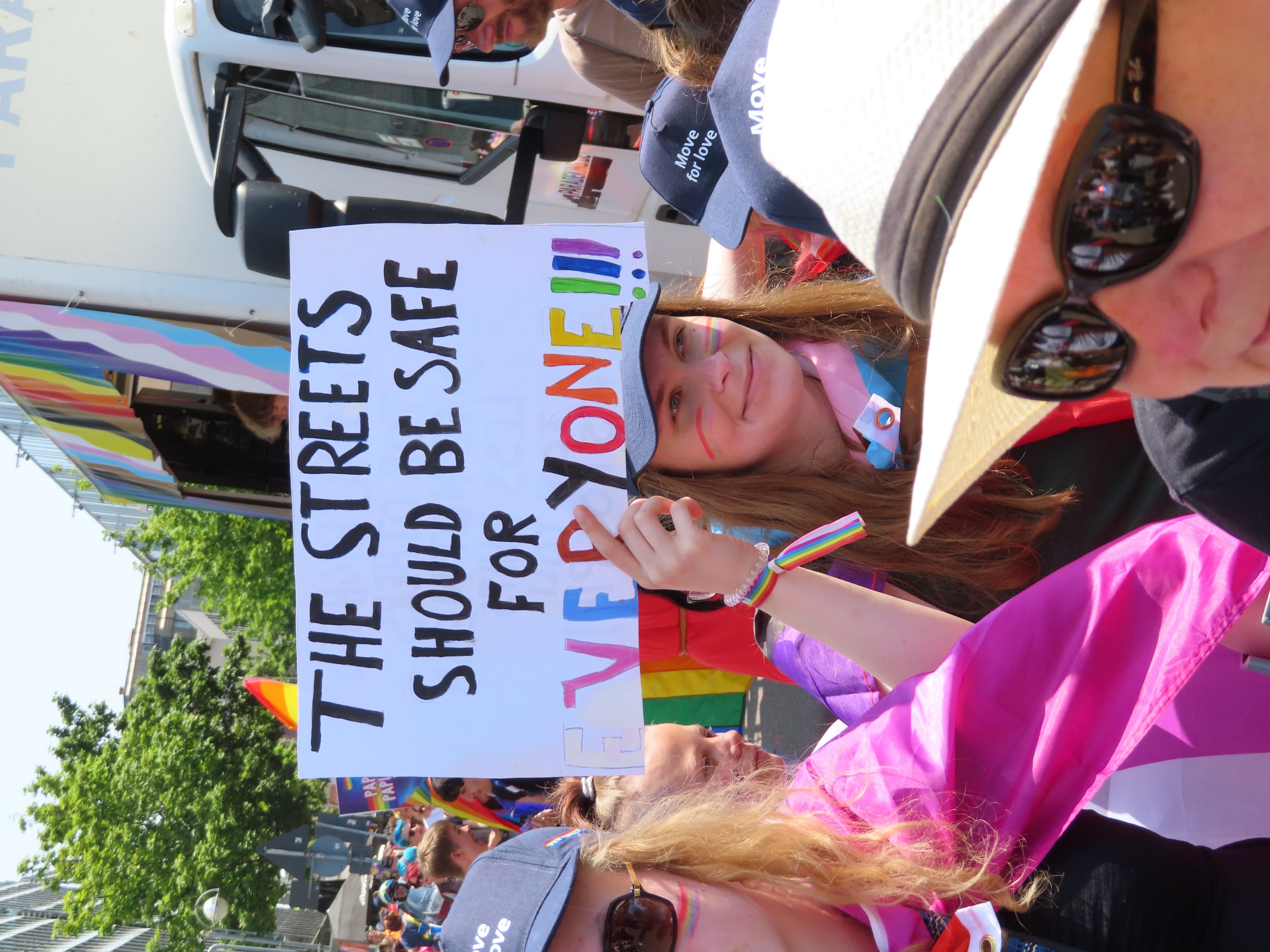 Eine Gruppe von Menschen steht vor einem Lastwagen und hält ein Schild mit der Aufschrift "Die Straßen sollten sicher sein", einige tragen Mützen und Schutzbrillen, andere halten Schilder, vor einem Hintergrund aus Bäumen, Gebäuden und einem klaren blauen Himmel.